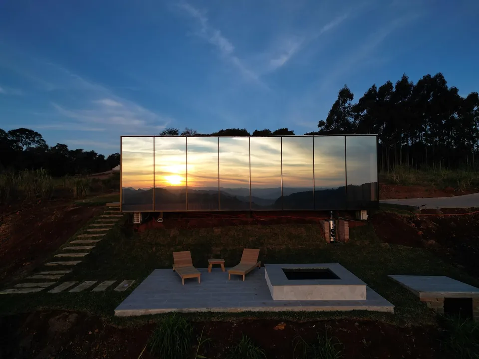 A parede de vidro espelhando o pôr do sol, com a fogueira e as espreguiçadeiras no deck