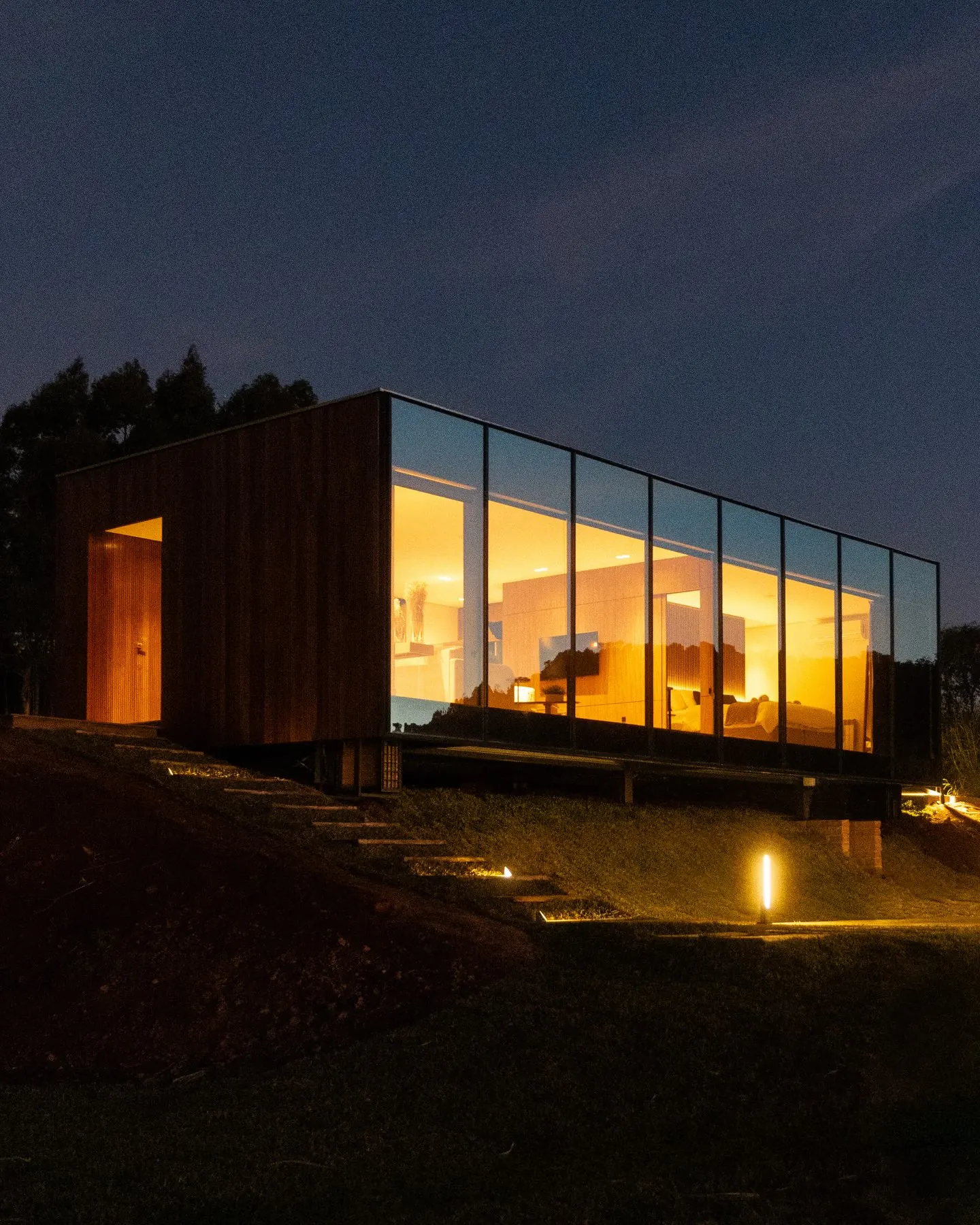 A Casa de Vidro Felix Hills acesa à noite, com a luz quente atravessando as paredes de vidro